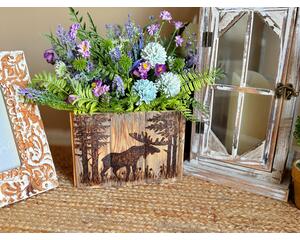 Image shows a wooden vase with a moose in the woods burned on the front. Box displays a faux wildflower arrangement with purple flowers. 